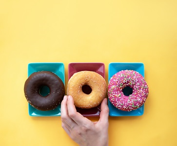 Drei Donuts auf bunten Tellern, Hand greift nach dem mittleren Donut auf gelbem Hintergrund.