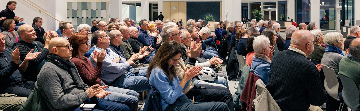 Publikumsversammlung: viele Personen sitzen in Räumen, klatschen, einige tragen Masken/Taschen.