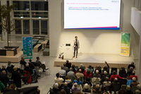 Redner steht hinter weißem Pult, im Hintergrund ist ein Bildschirm mit einer Präsentation