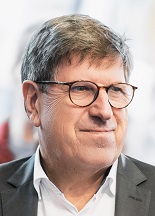 Mann mit grauen Haaren, Brille und Anzug in einer Innenraumszene.