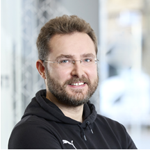 Person mit dunklem Hoodie, Gesichtsunsichtbar durch Unschärfe, stehend im Innenraum.