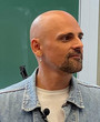Ein Mann mit kahlem Kopf, grauer Hoodie und Denim-Jacke, vor grünem Hintergrund.