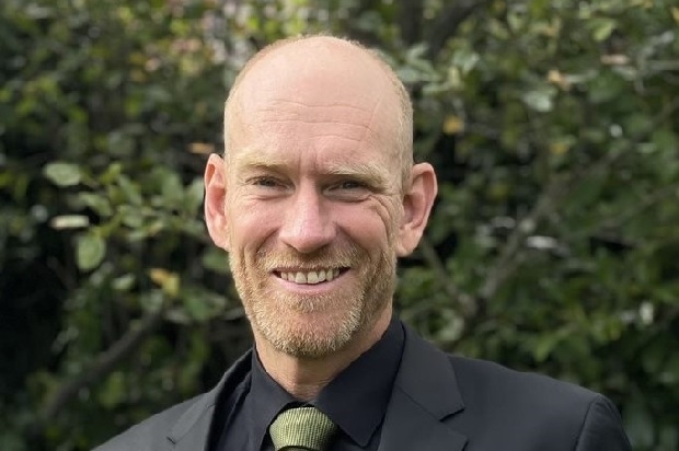 Ein Mann in Anzug mit grünem Krawattenband vor grüner Bürowiese (Blende über Gesicht).